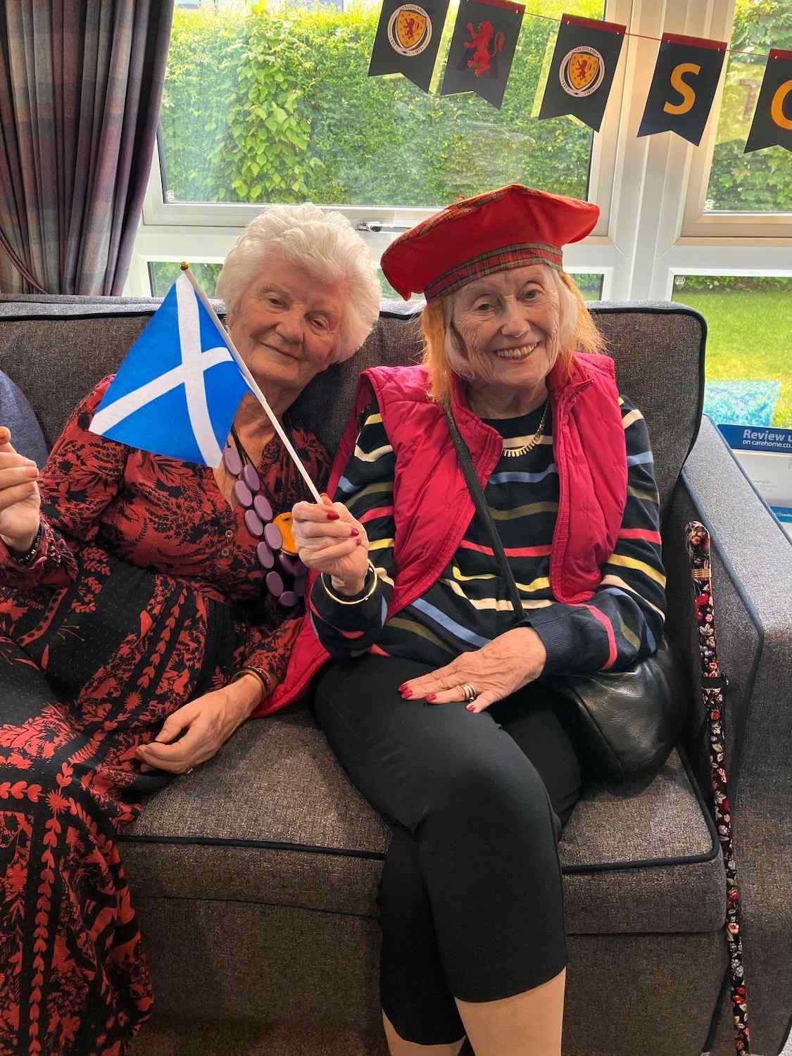 Two elderly women sit smiling waving a Scotland flag